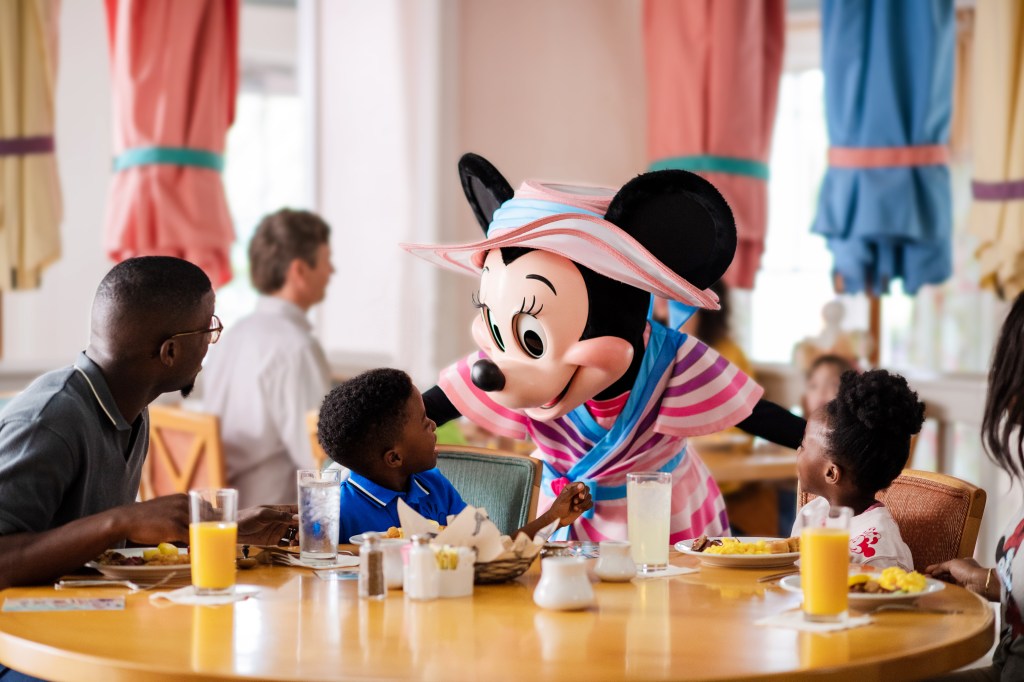 Minnie Mouse greets a young boy at a hotel breakfast. She is wearing a dress with purple, magenta, and light pink stripes, with blue accents. 