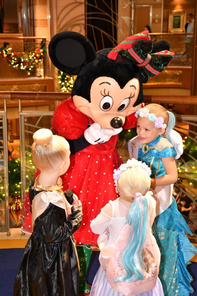 Three small girls wearing princess dresses hugging Minnie Mouse, dressed in Christmas attire.