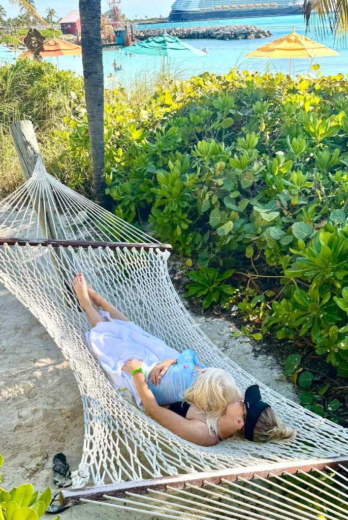 Mother lying with daughter in hammock on Disney's Castaway Cay, with Disney cruise ship in background.